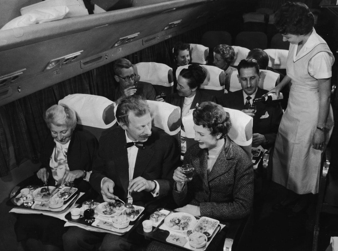 In the 1950s, an interior view of a commercial passenger plane shows a flight attendant pouring a glass of wine for a man who sits next to a couple who are toasting each other with full glasses.