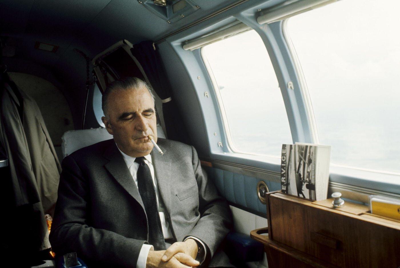 Georges Pompidou smoking a cigarette with his eyes closed, sitting near a window on an airplane during his 1969 presidential campaign.