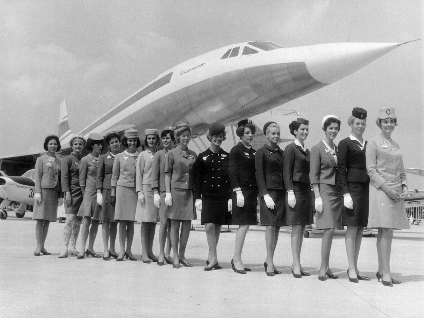 A lineup of air stewardesses from different airlines attending to passengers on board the supersonic jet Concorde, standing in front of a scale model of the aircraft.