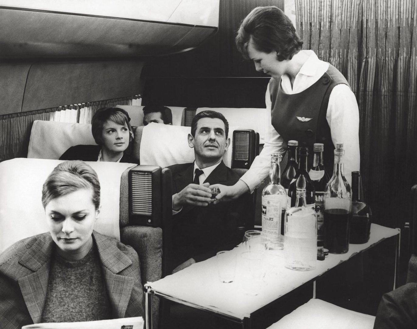 Air hostess offering drinks to passengers on board plane. 1960s.