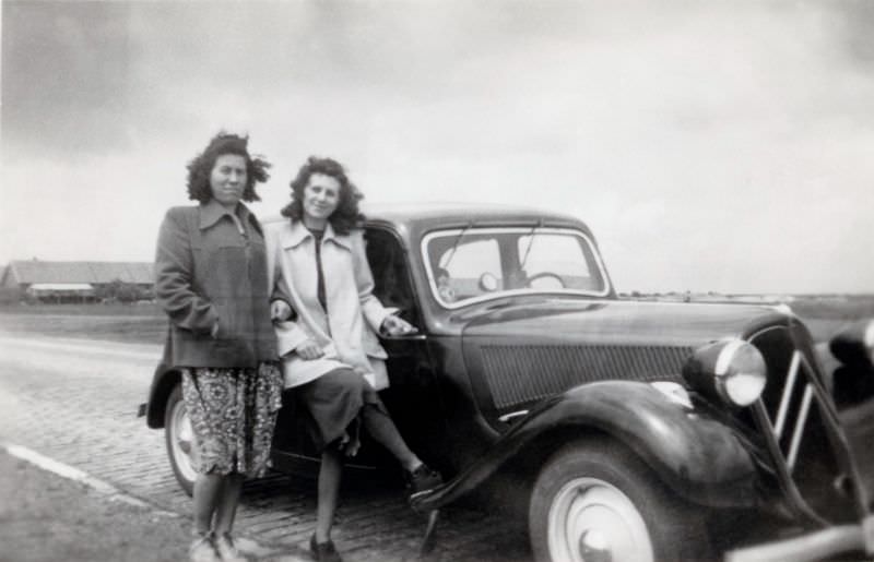 Two middle-aged ladies with Citroën 11 CV on cobbled road in countryside, 1950