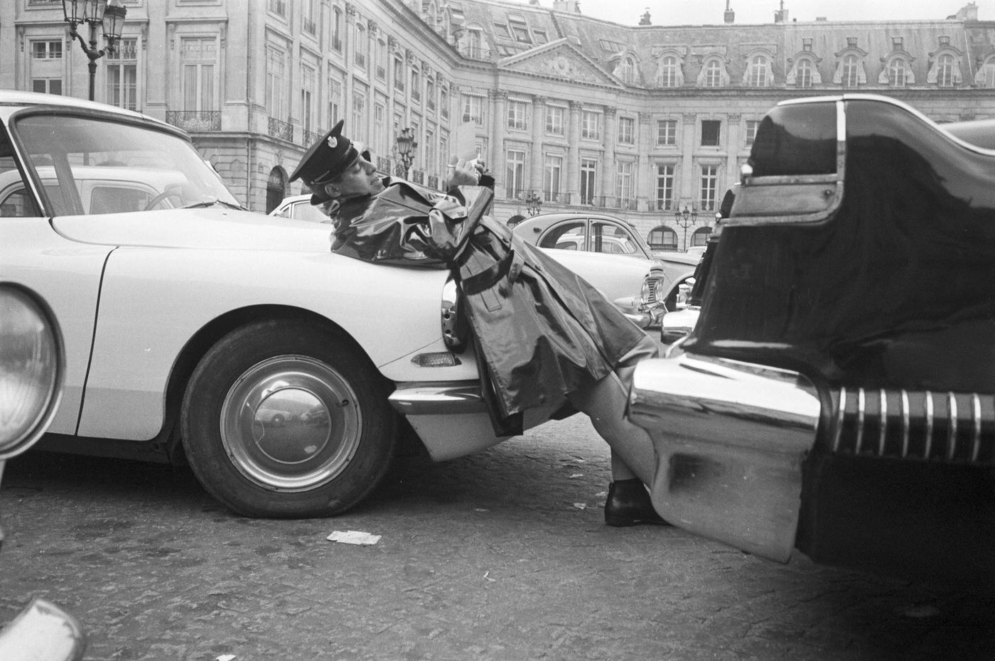 Jerry Lewis filling out a fake parking ticket while lying on the hood of a black Citroën DS on Place Vendôme in Paris, 1965