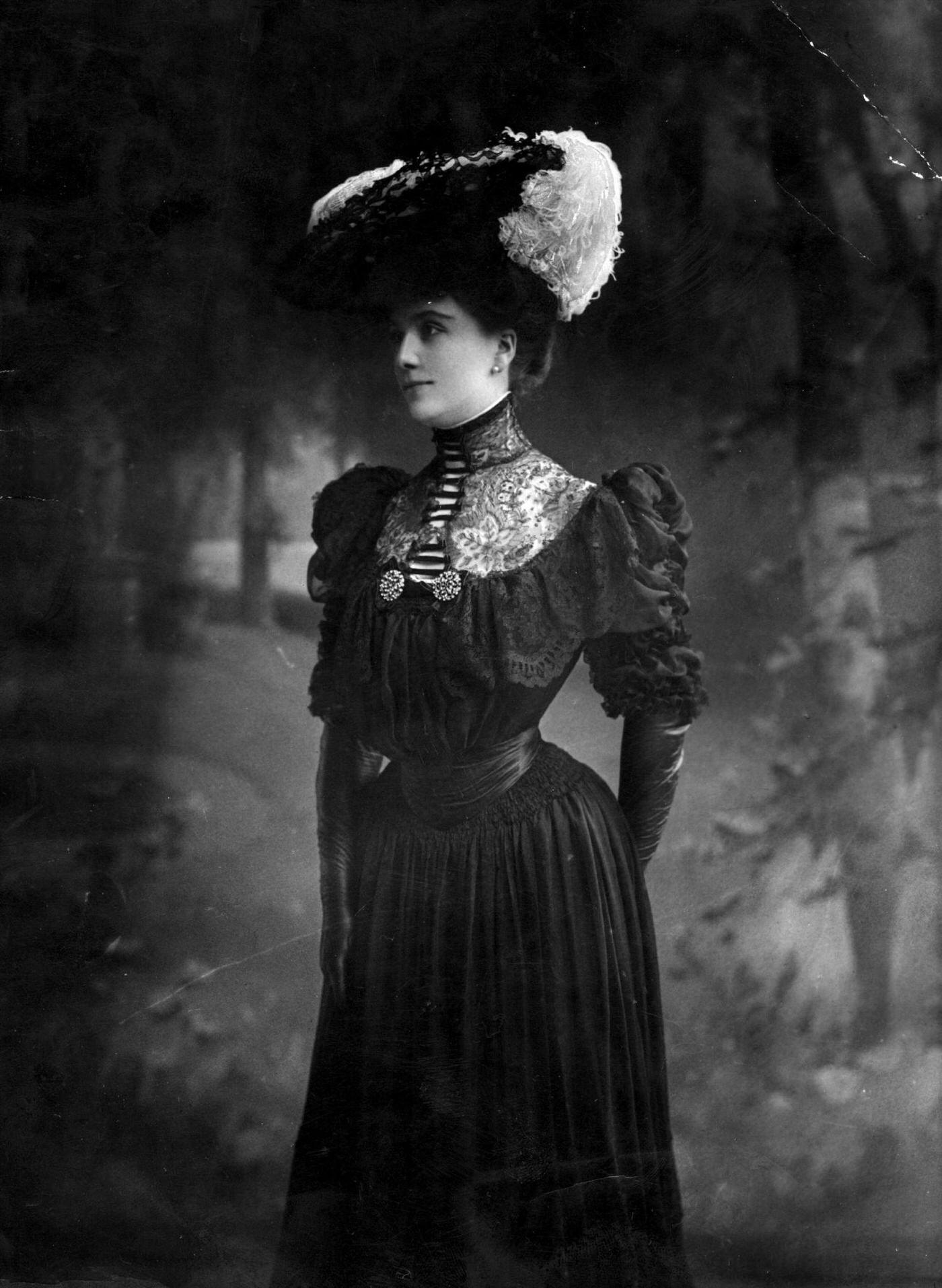 Miss Lewis wears a dark period dress with alluring lace detail at the throat, set off beautifully by a feather-trimmed hat