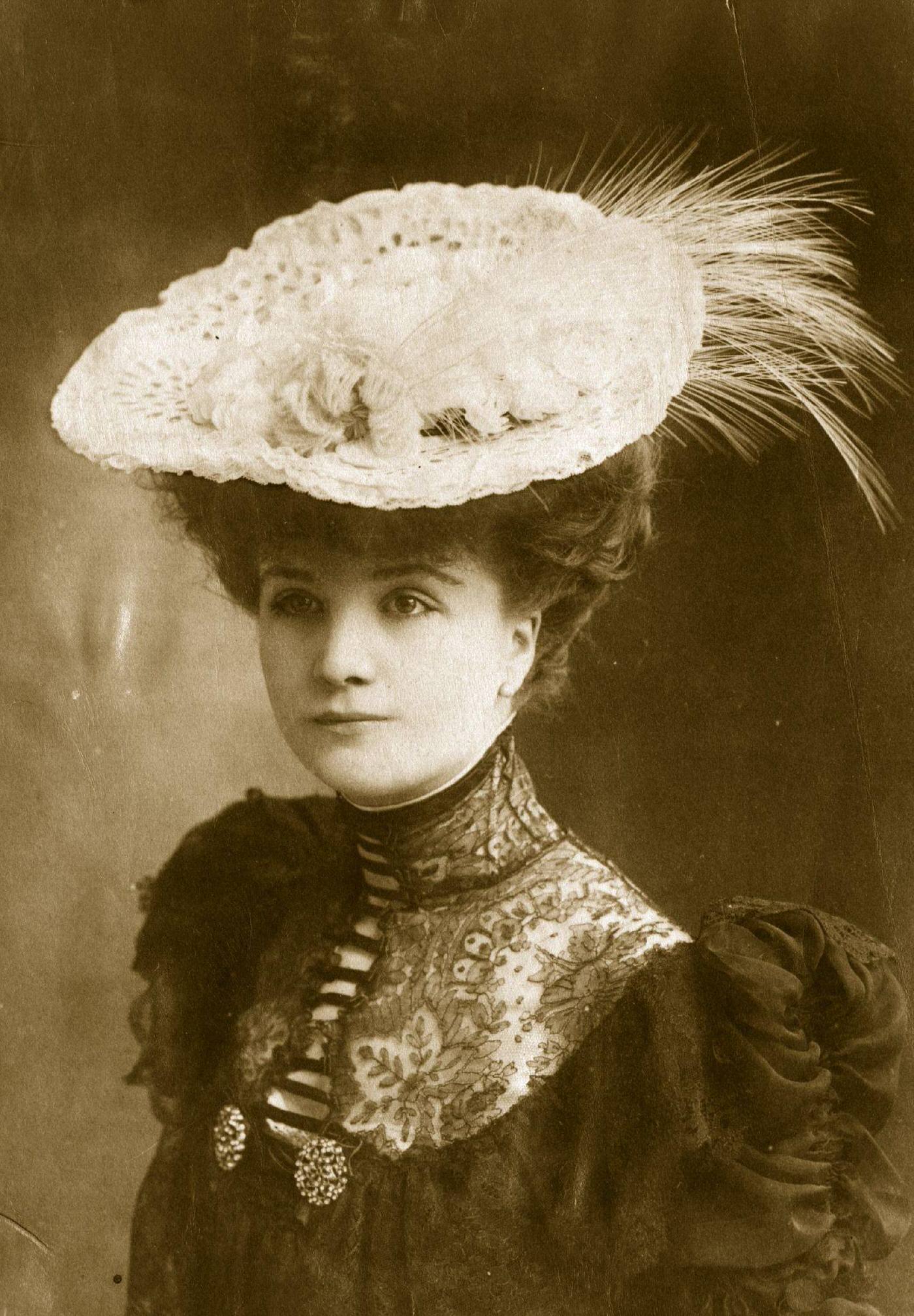 Miss Lewis wears a dark period dress with alluring lace detail at the throat, set off beautifully by a feather-trimmed hat