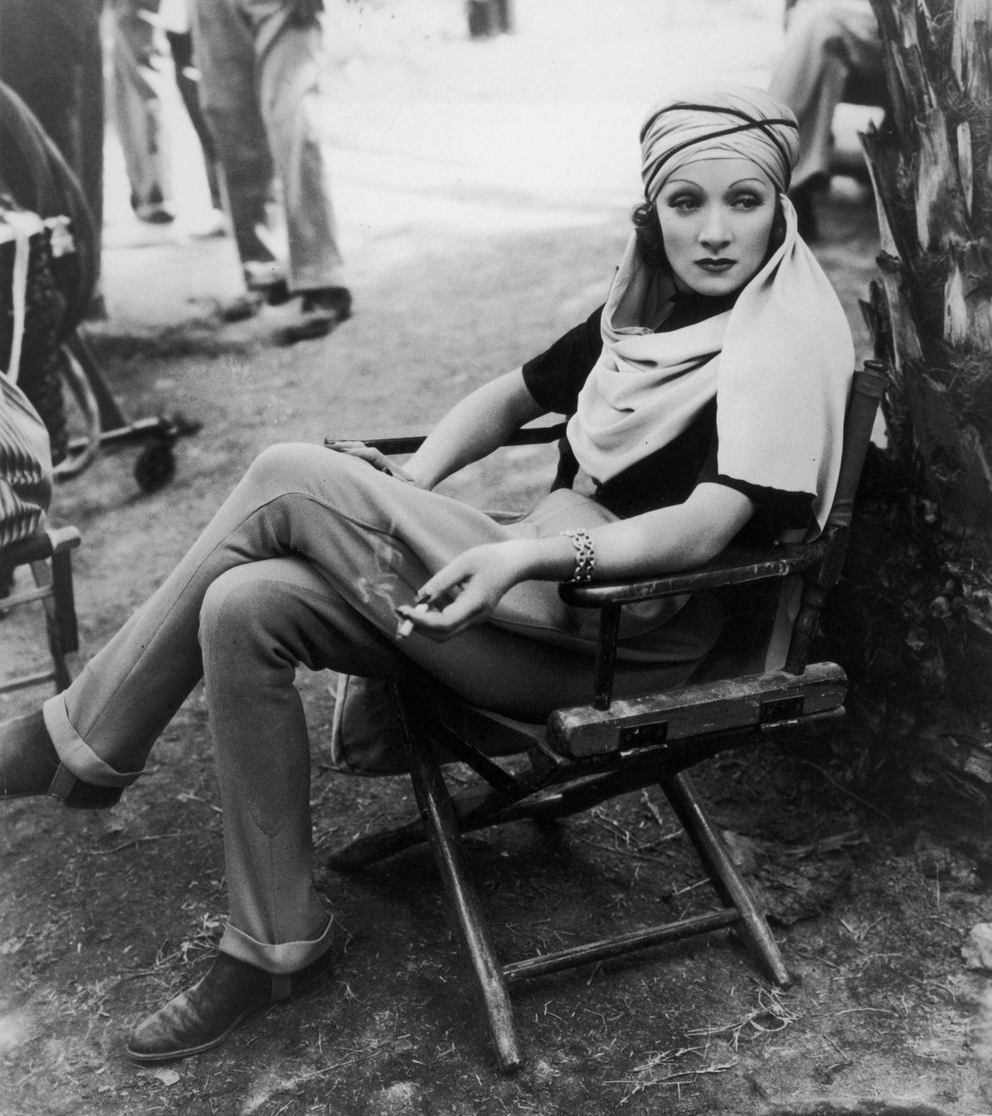 Marlene Dietrich sits in a canvas chair, wearing a turban, and holding a cigarette on the desert location set in 1936.