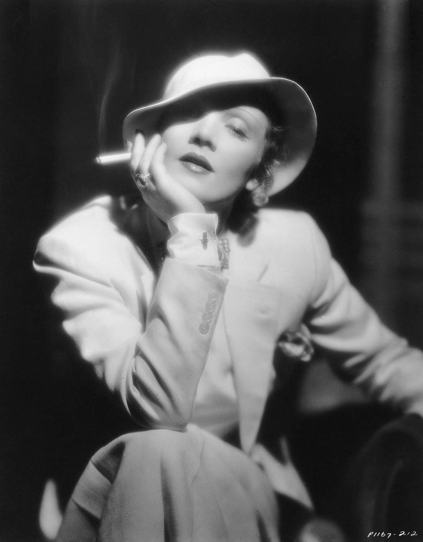 A photograph of a German-born Marlene Dietrich holding a cigarette, dressed in a tailored suit, hat, bow-tie, and cufflinks.