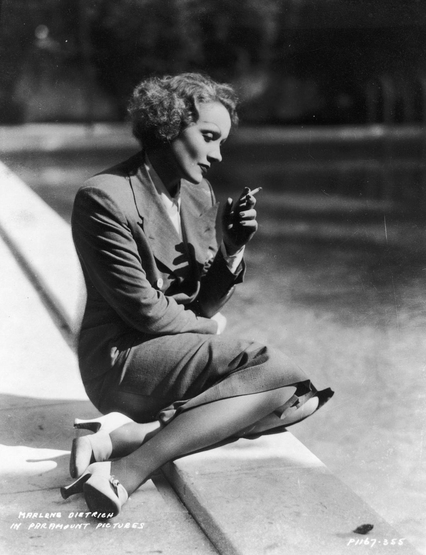 In 1932, Marlene Dietrich kneels by a lake while smoking a cigarette in a double-breasted suit.