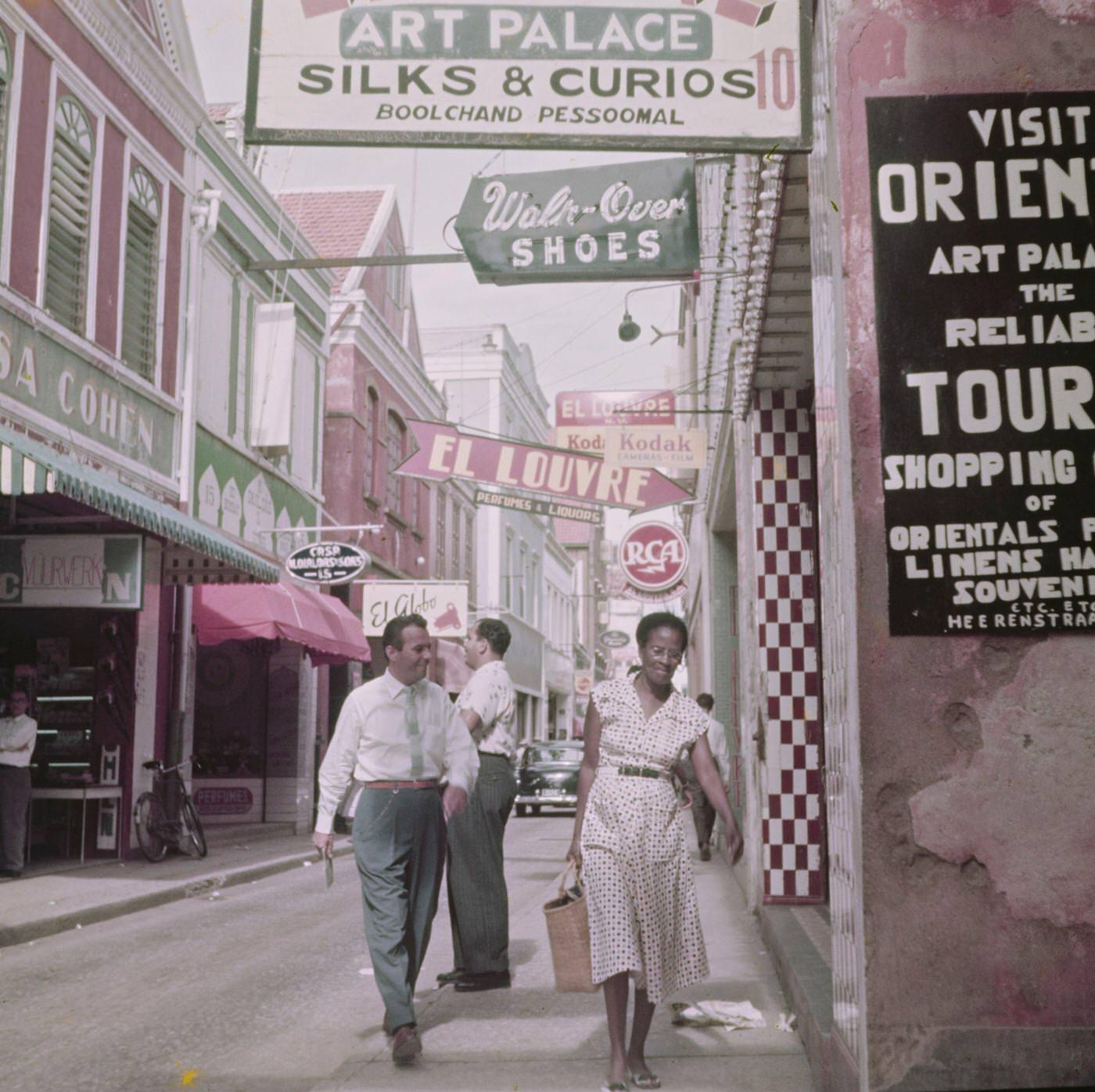 Pedestrians and residents walking along a shopping street in Willemstad, capital city of the island of Curaçao, part of the Kingdom of the Netherlands in the southern Caribbean Sea, 1955.