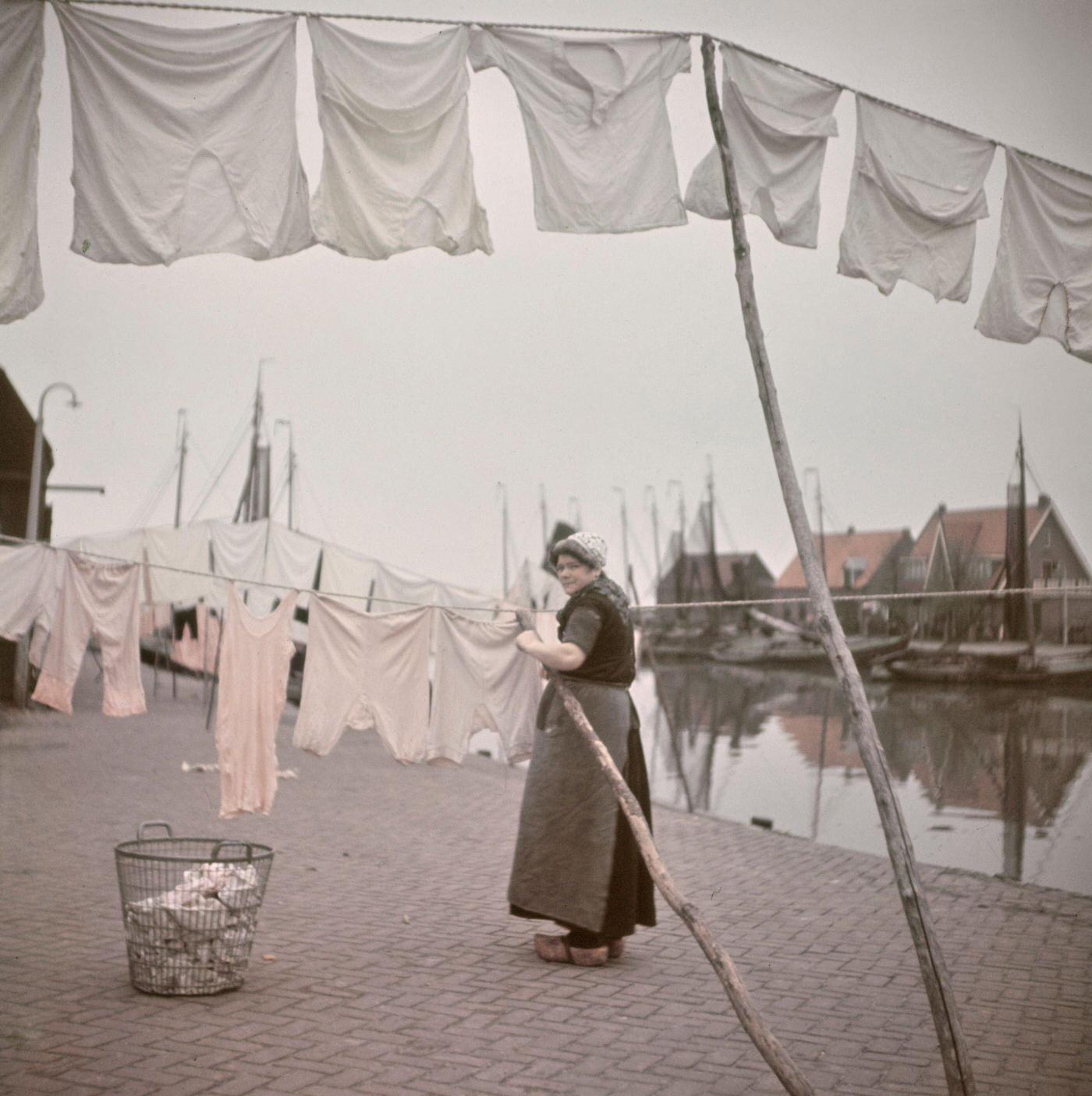 A woman wearing traditional clogs (klompen) hangs out washing to dry on lines on the quayside beside fishing boats moored in the harbour of the village of Spakenburg in the province of Utrecht, Netherlands, 1950.