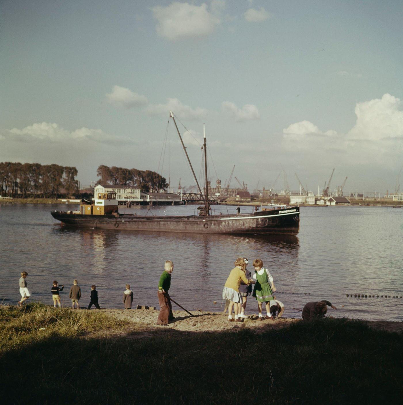 A barge passes children playing on a canal bank in a suburb of Amsterdam, capital city of the Netherlands, 1950.