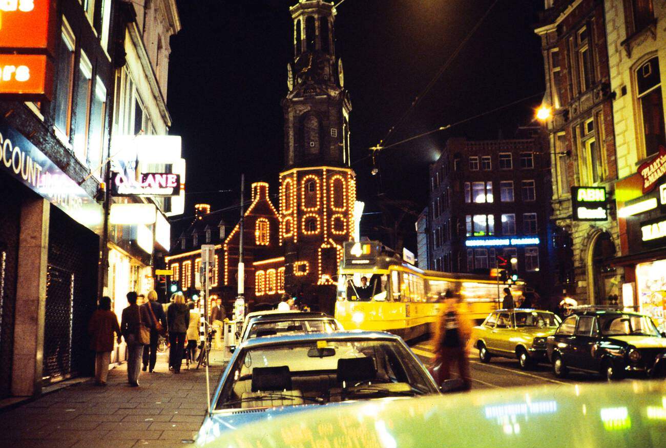 Neon lights at night in the city center of Amsterdam, The Netherlands, during the 1970s-1980s. A yellow tramcar and Munttoren tower are visible.