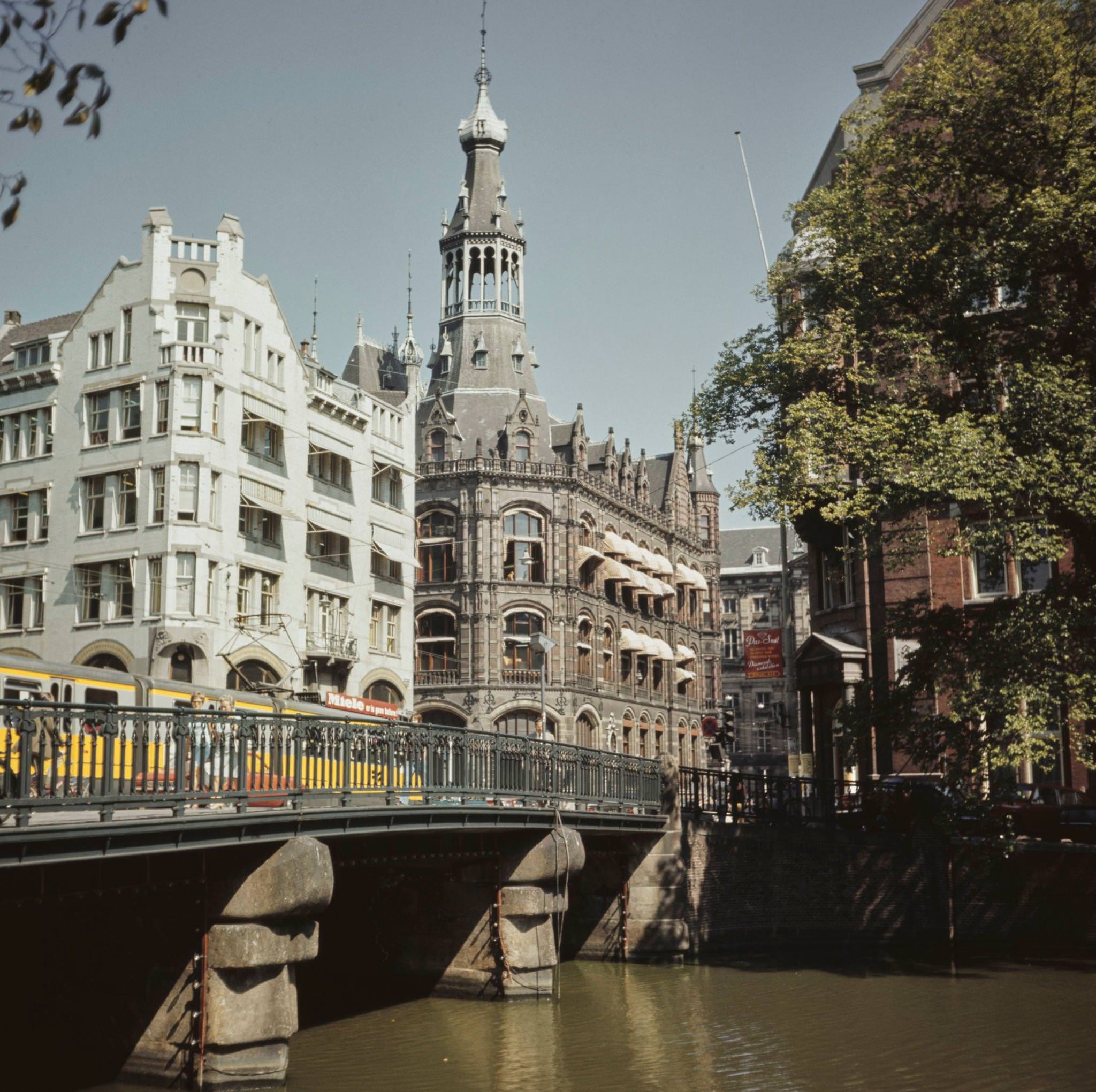 A tram runs along Raadhuisstraat on a bridge over Singel Canal in front of the Central Bank and Old General Post Office in the center of Amsterdam, capital city of the Netherlands, 1970