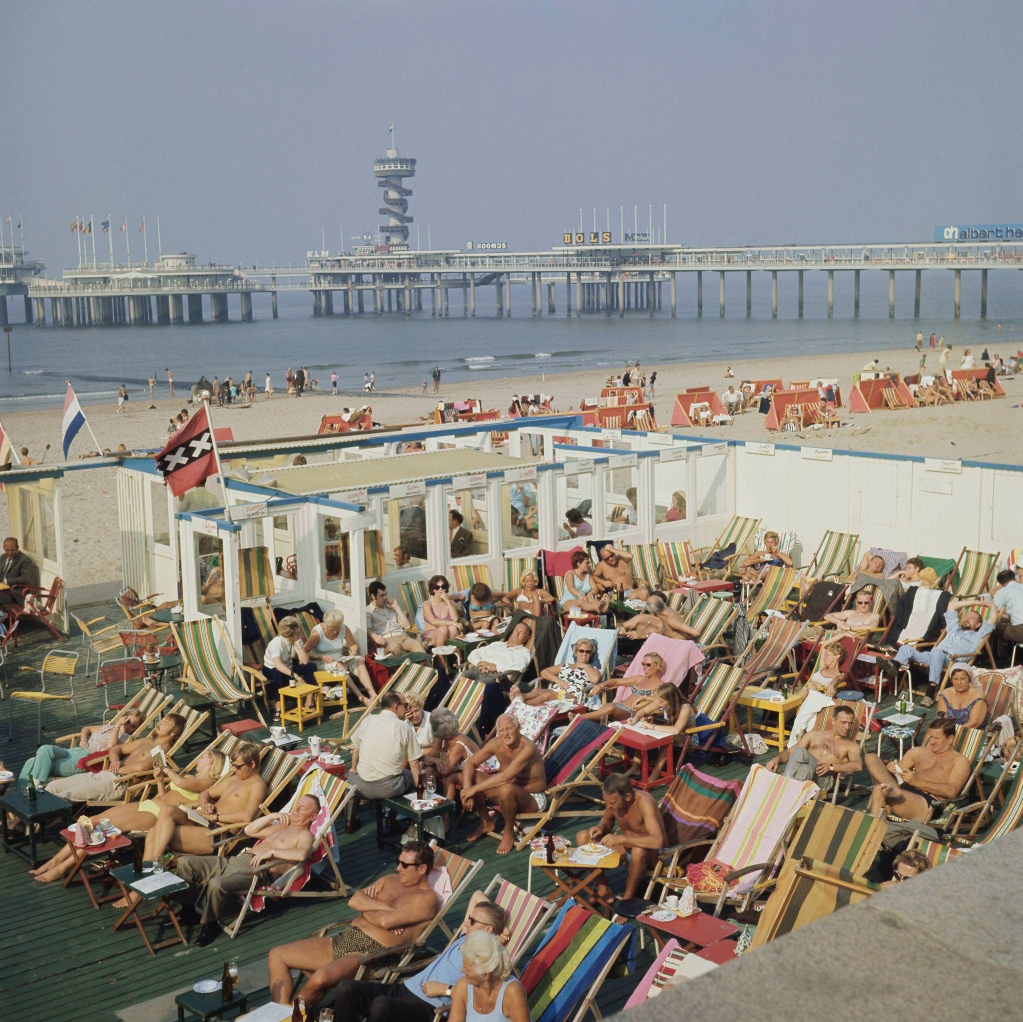 Visitors and holidaymakers sunbathe on deckchairs on a terrace beside the sandy beach at the seaside resort of Scheveningen near The Hague in the Netherlands, 1970s