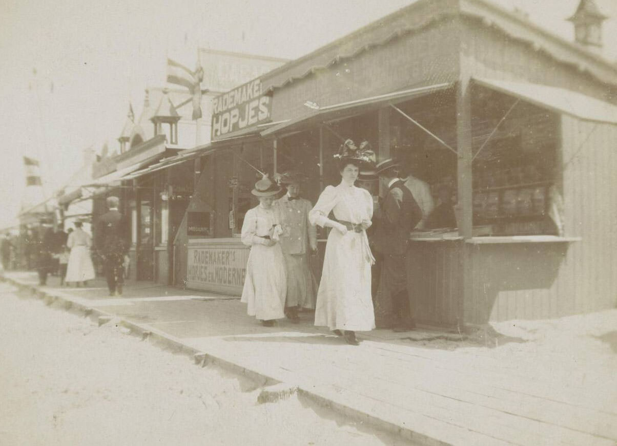 Beach scene with walkers on planks past stalls, 1900s
