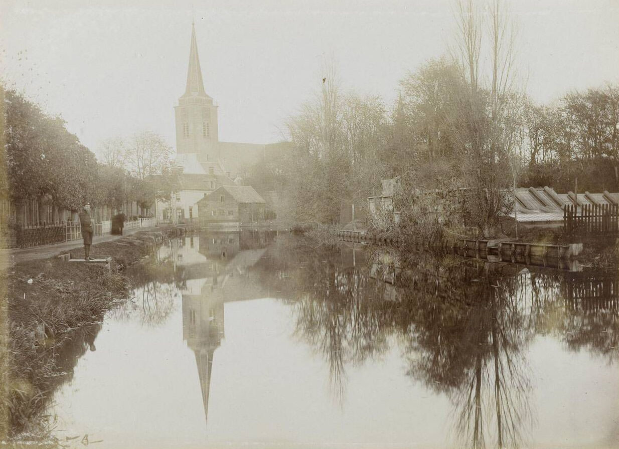 View of a wide ditch with a road along it, and a church on the background, Netherlands, 1900
