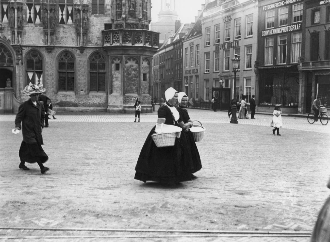Pair of Dutch women carrying baskets crossing a town square, Netherlands, 1900