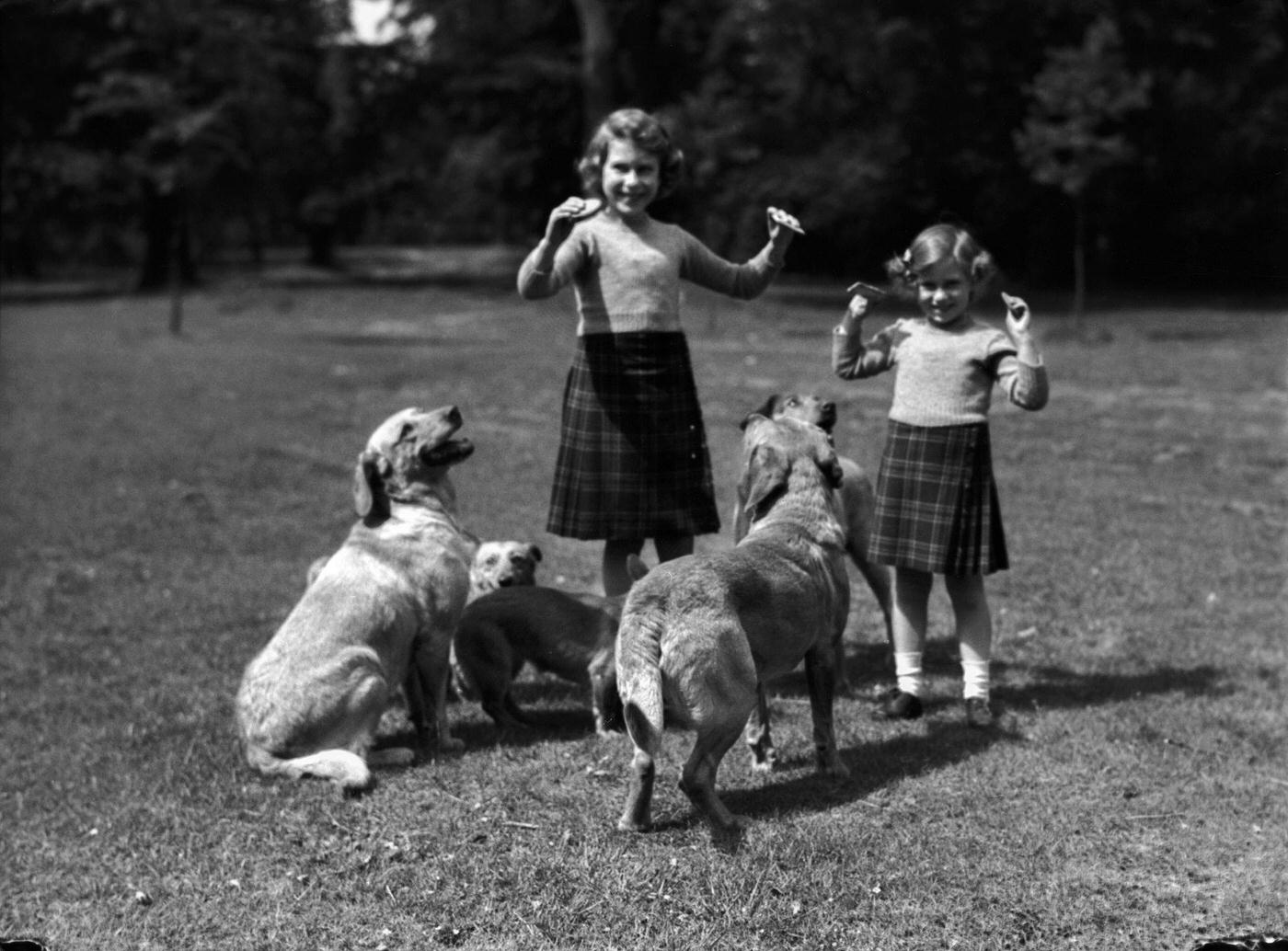 Princess Elizabeth and her sister Princess Margaret wearing kilts and feeding biscuits to dogs at the Royal Lodge, Windsor, UK.