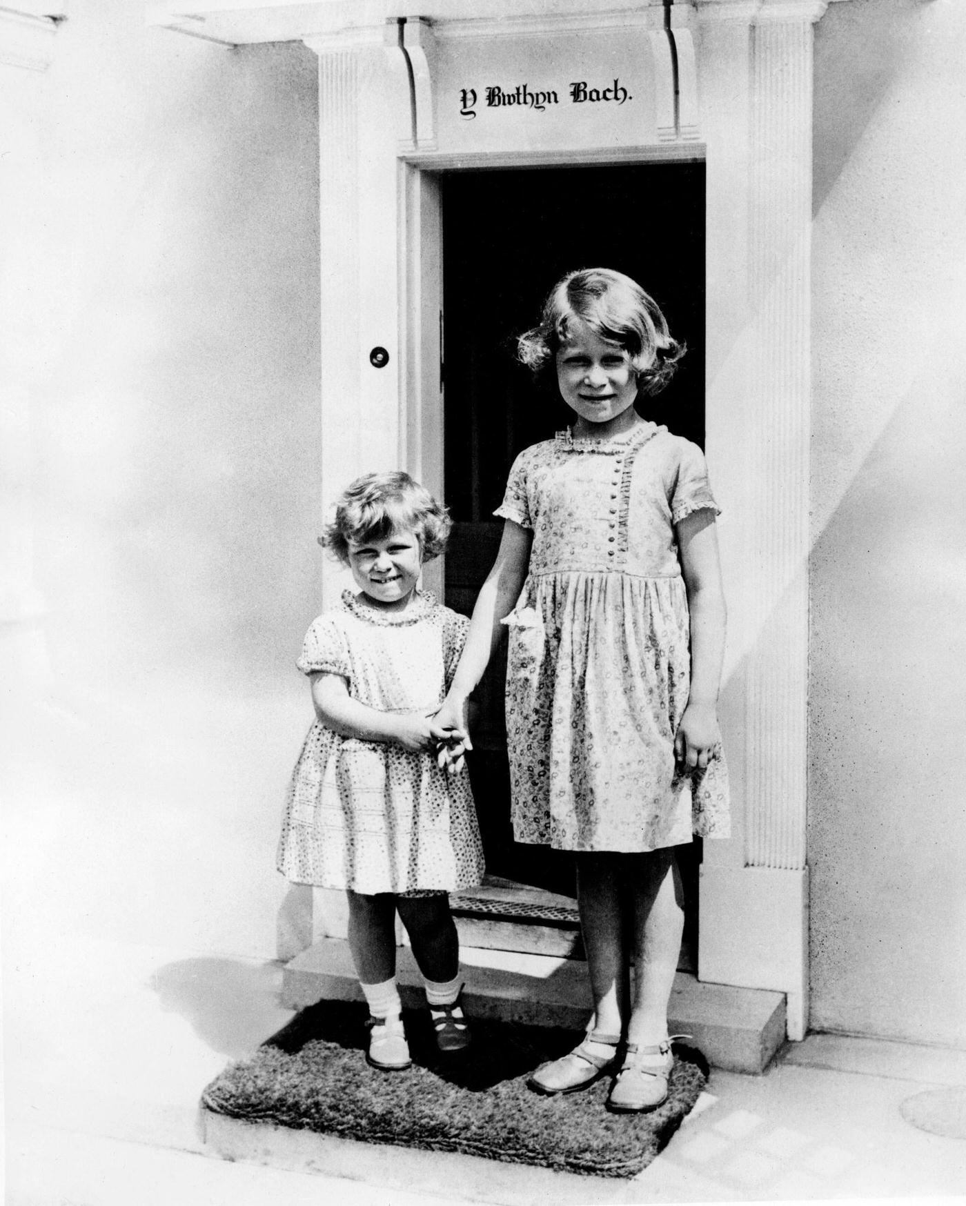 Princesses Margaret and Elizabeth (the future Queen Elizabeth II) posing in the doorway of a miniature Welsh cottage.