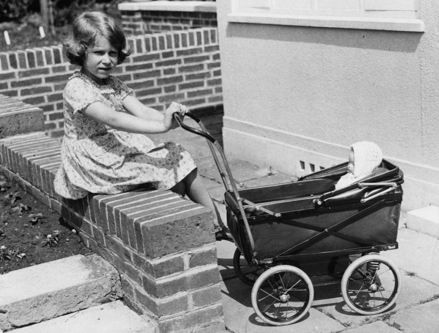 A childhood photograph from 1933 shows Queen Elizabeth II with a toy doll in a pram, in the grounds of the Royal Lodge at Windsor.