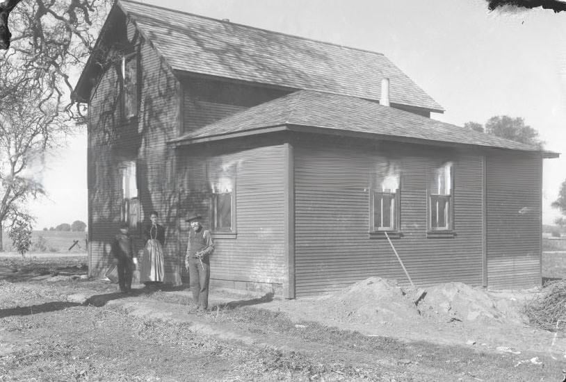 View of three people standing in front of a house, 1890