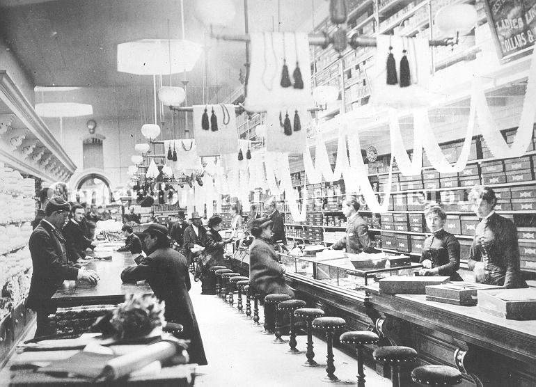 Sales people wait on customers seated at counter in shirt department, 1890