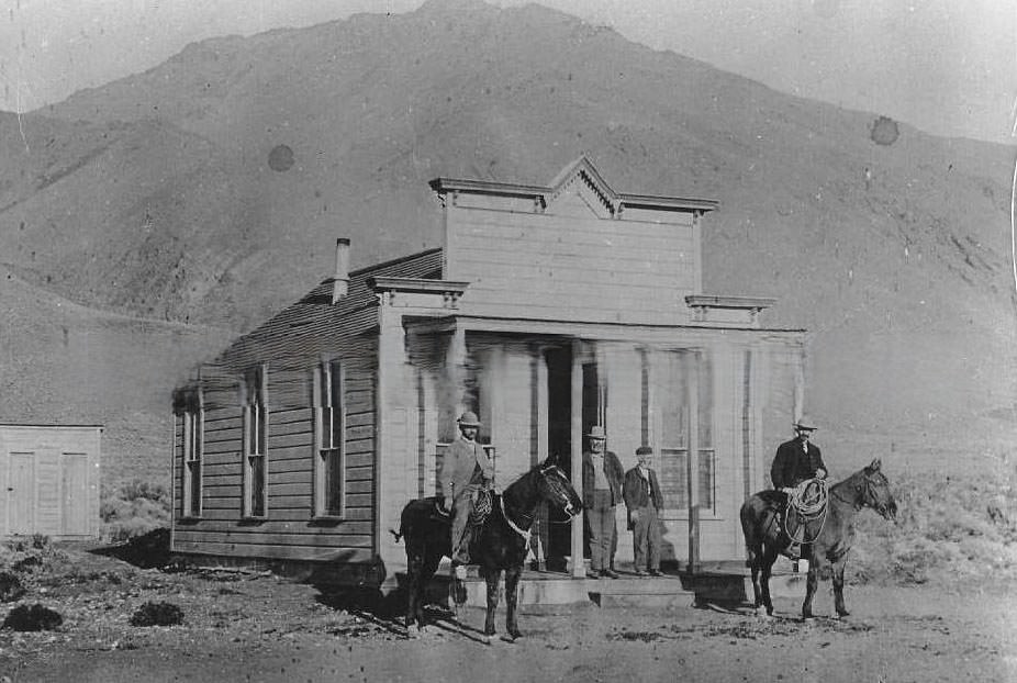 Two men mounted on horses and two men standing on the porch of a house, 1890