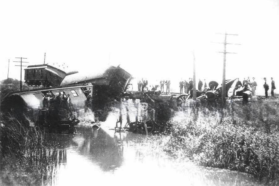 Train wreck over a slough in Sacramento. Groups of men standing on the bank of the slough, bridge and on the wrecked cars and locomotive, 1890