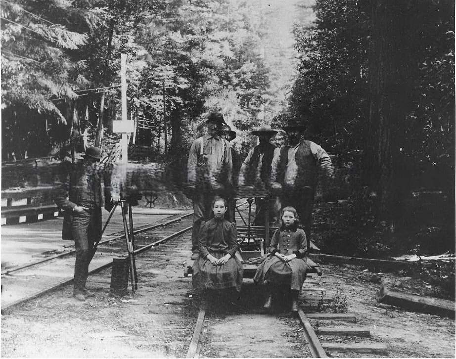 Four men stand on a railroad handcar, two school-aged girls sit on front of it, 1895