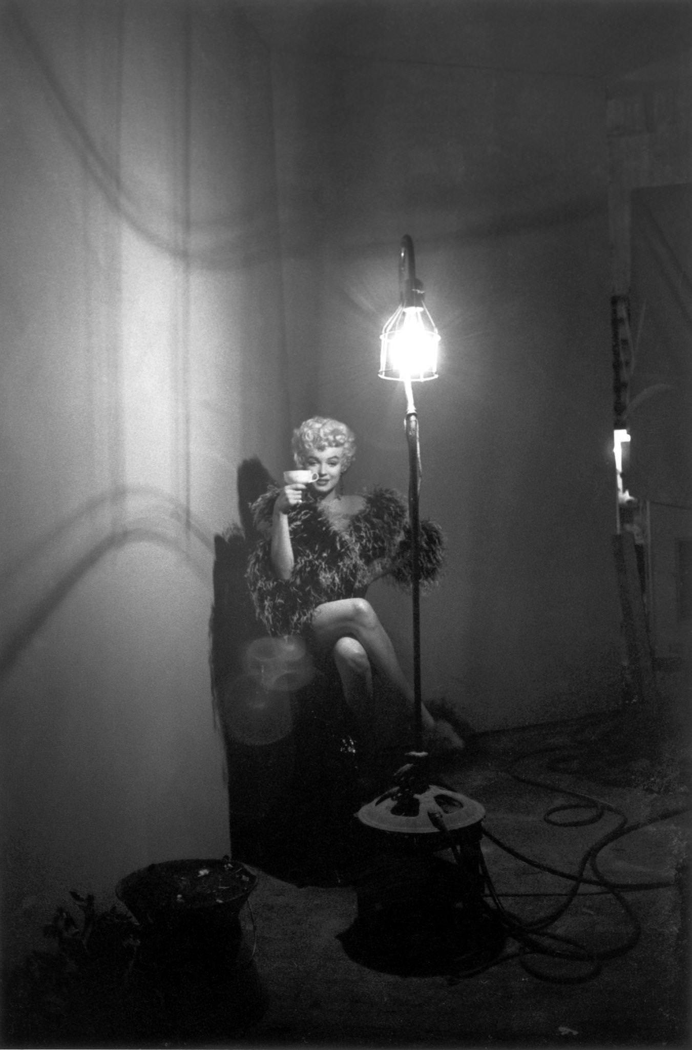 Marilyn Monroe in a feather boa holding a teacup as she sits next to a tall floor lamp in 1954 during the filming of "The Seven Year Itch" in Los Angeles