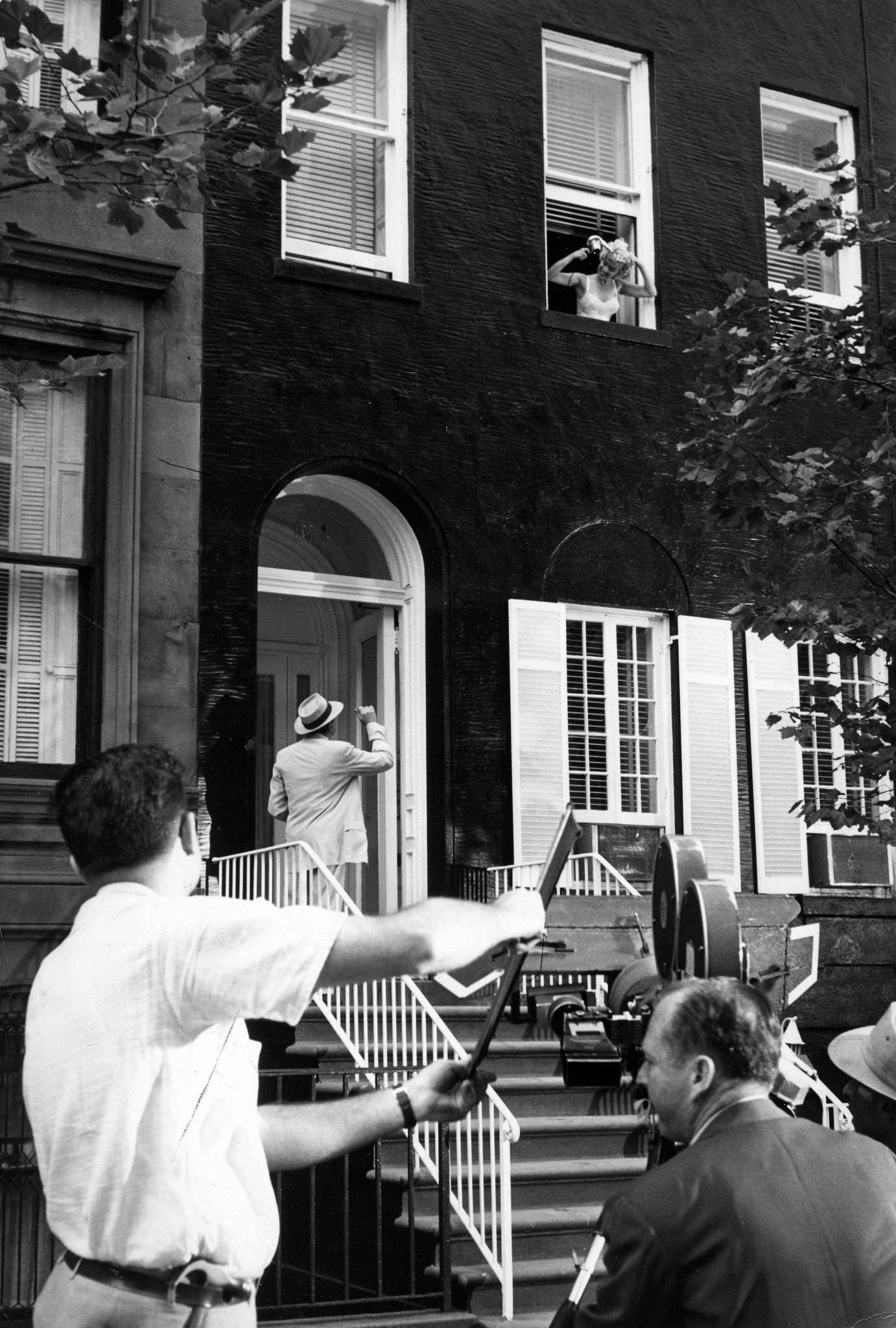 Marilyn Monroe leans out of a window wearing a slip and holding a hairdryer with co-star Tom Ewell standing below