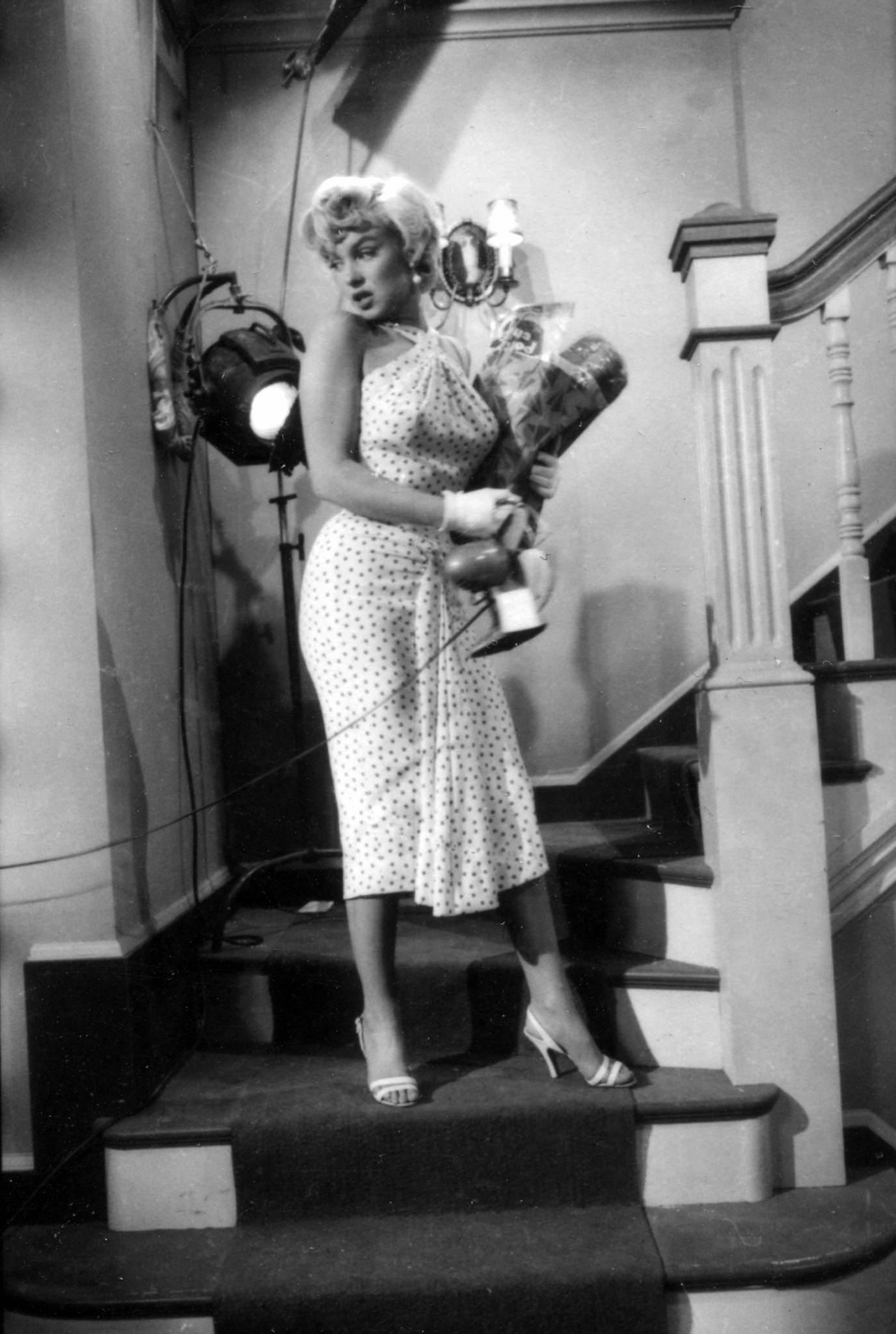 Marilyn Monroe, wearing a polka-dot dress, ascends a staircase holding a bag of groceries and a desk fan in her arms in 1954 during the filming of "The Seven Year Itch"