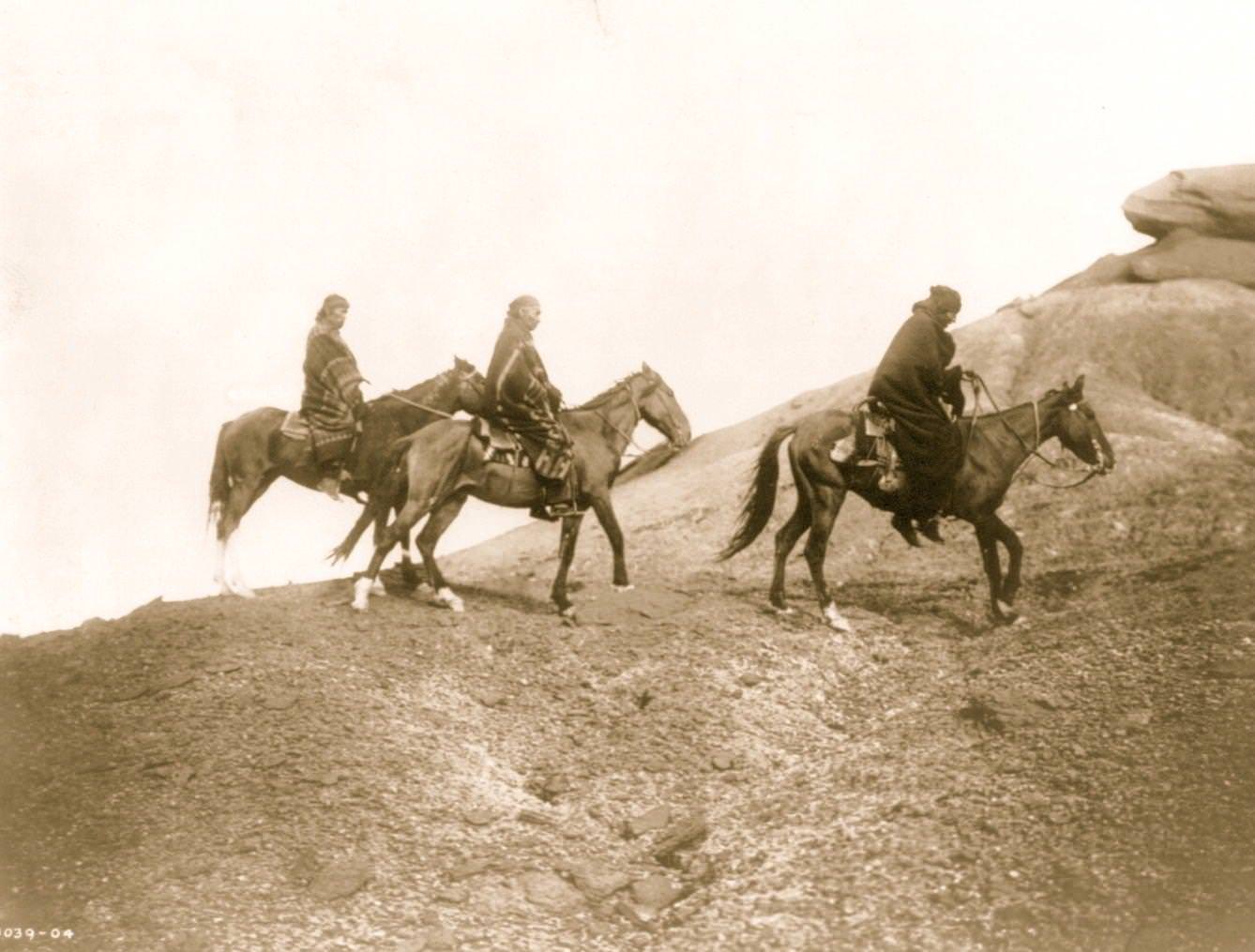 Three Navajo Indians on horseback make their way over hilly terrain, 1905