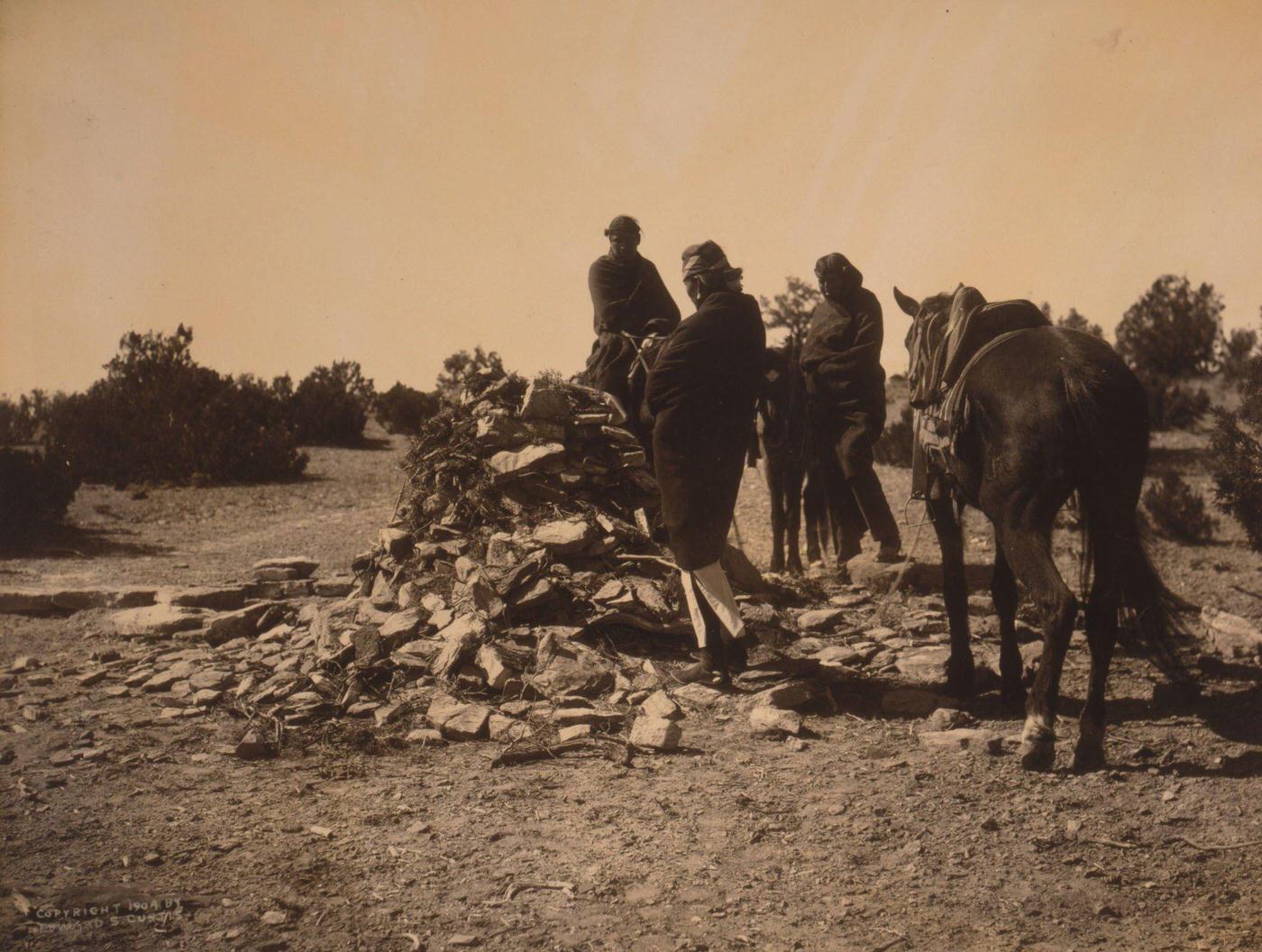 Three Navajos with horses, gathered before mound of rocks and vegetation, 1904