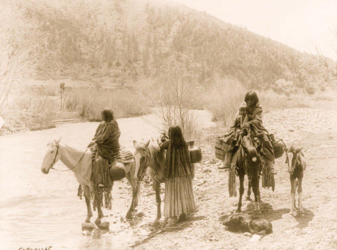 Three Apaches, one with a child, with horses laden with water jugs, a colt, and a dog stopped beside a stream, 1903