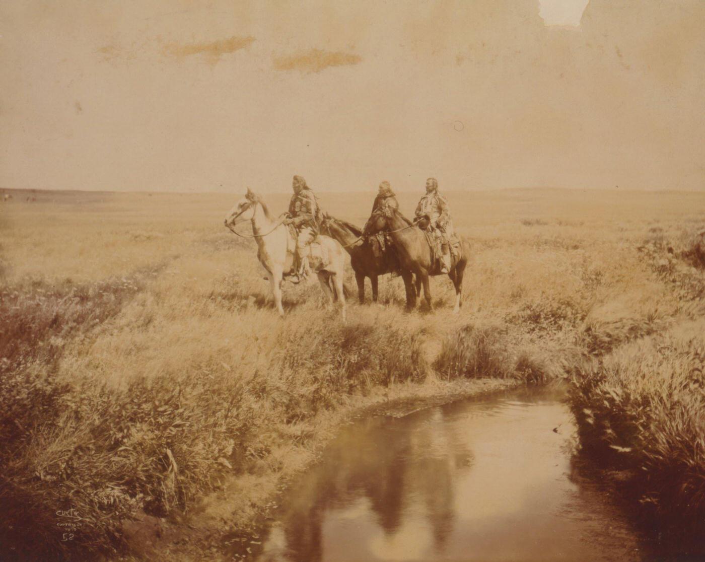 Three Piegan men on horseback in open grassland near pond, 1900