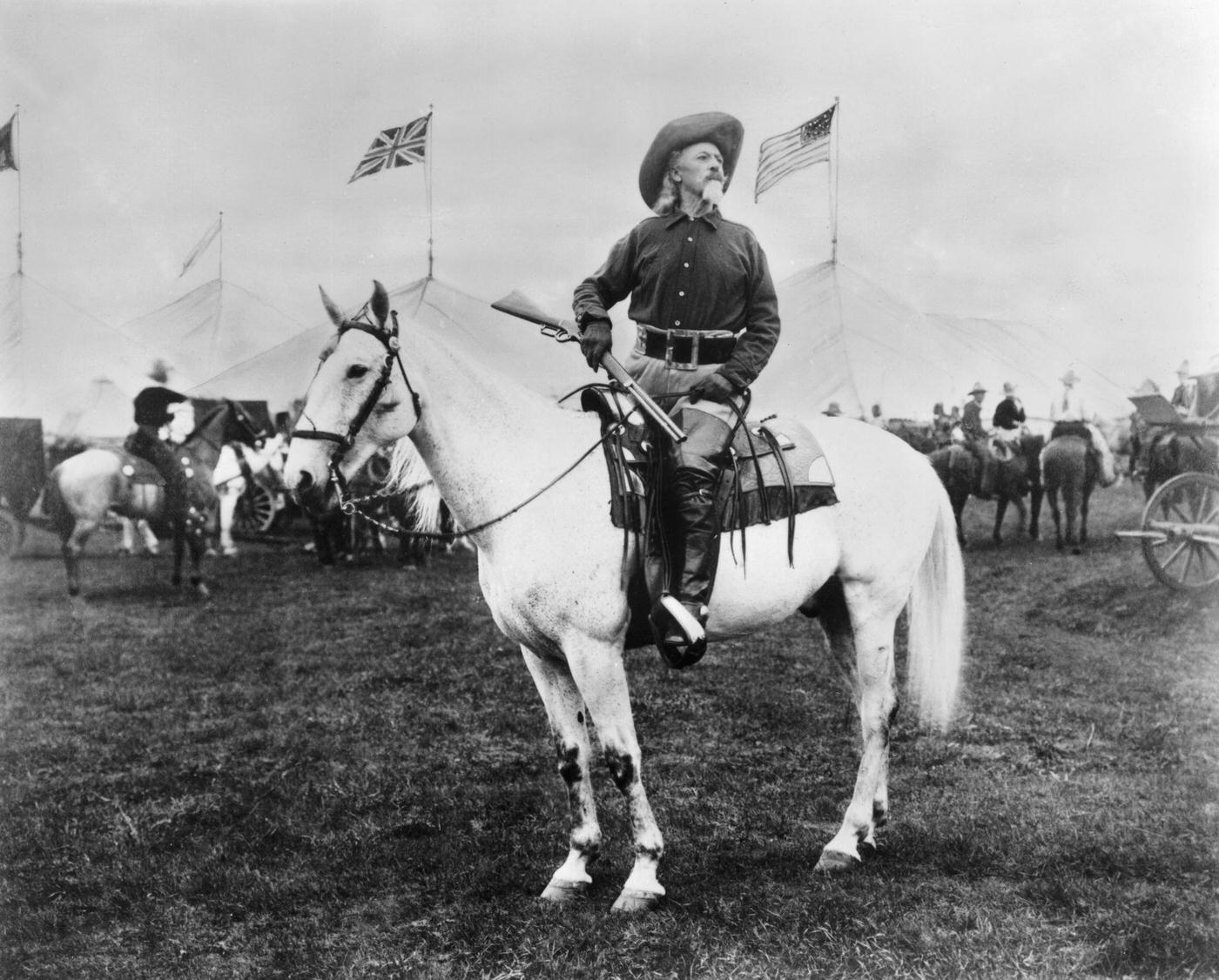 William 'Buffalo Bill' Cody (1846 - 1917) American entertainer, sitting on horseback and holding a rifle, looks off into the distance as British and American flags fly around him. Tents for his Wild West show are in the background.