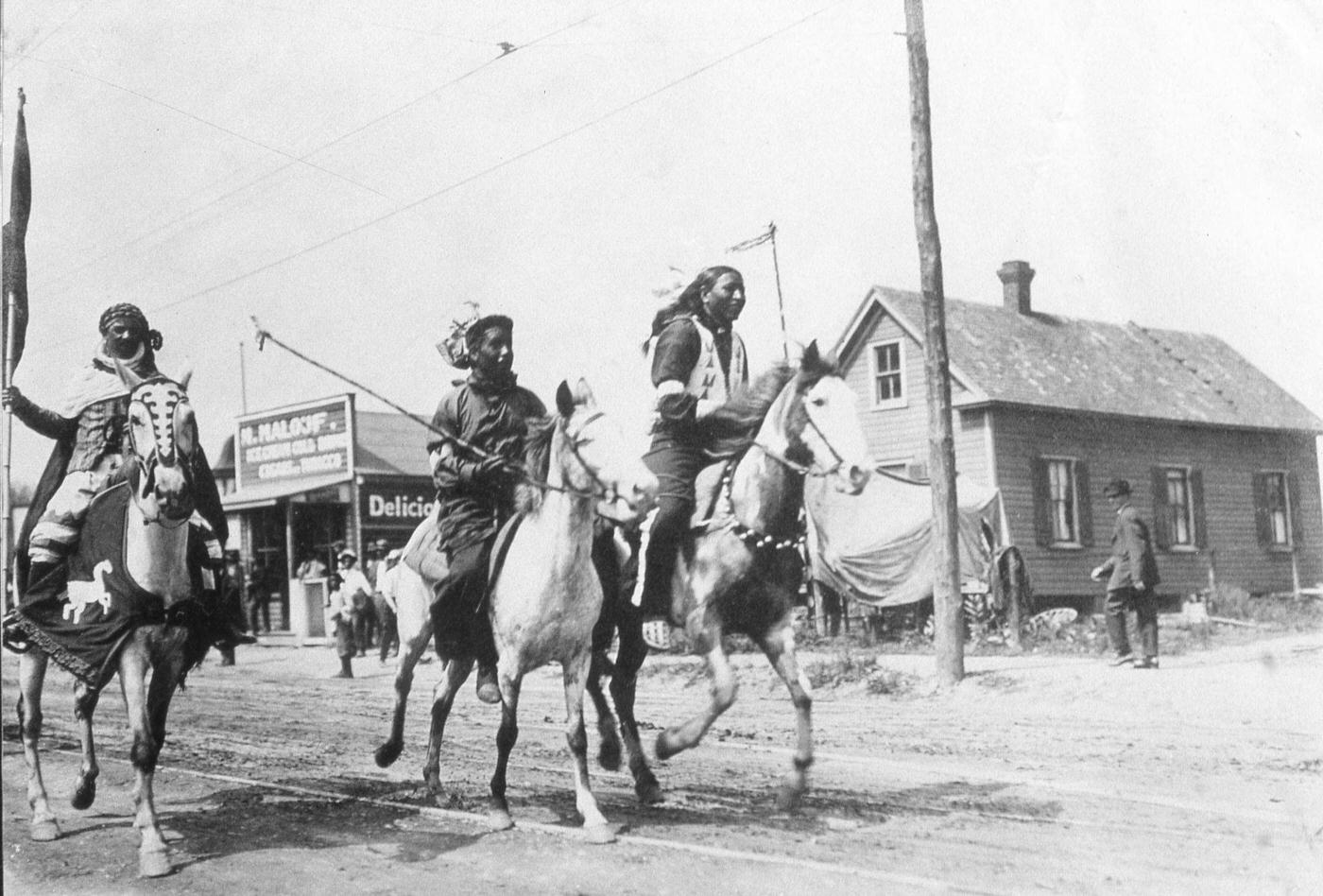A photograph shows three Native American men riding horses during Buffalo Bill's Wild West Show, United States, late 19th century. They race down a road in a rural town.