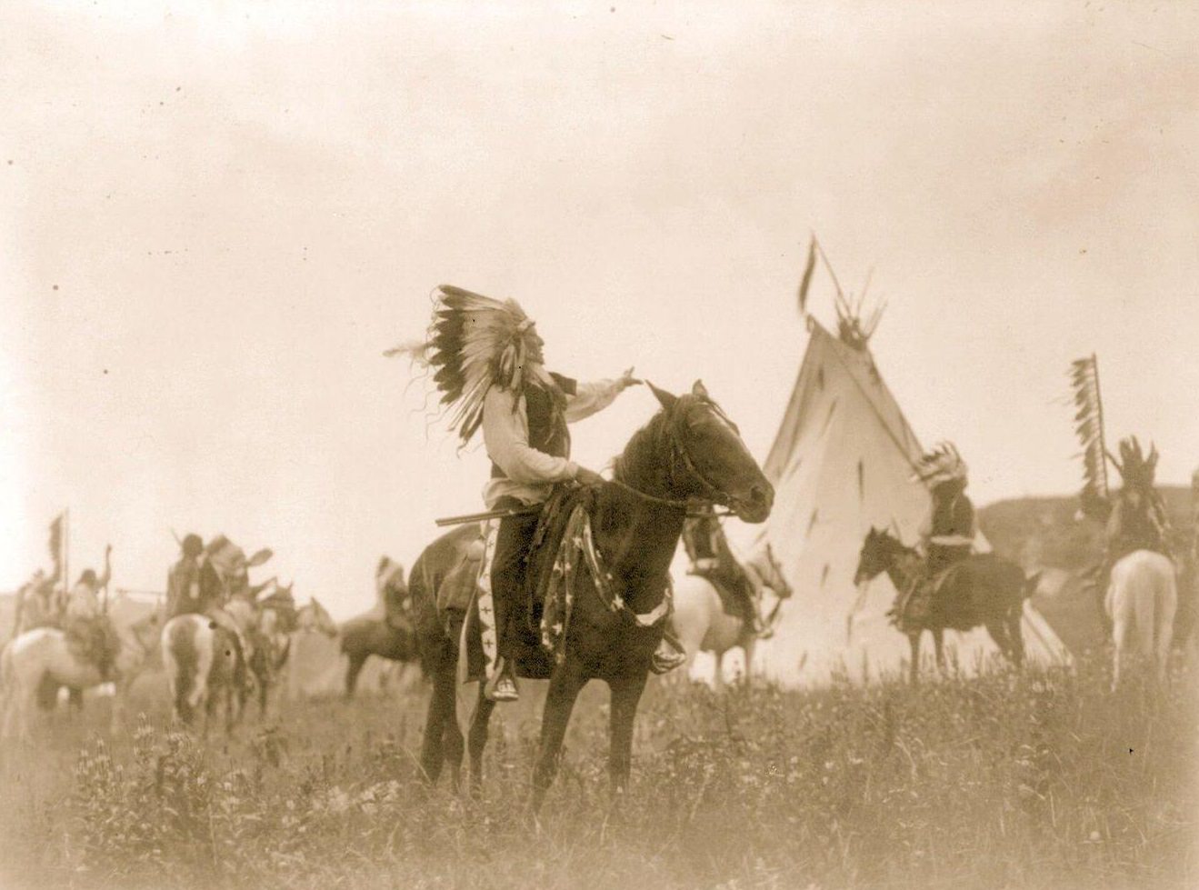 Dakota man, wearing war bonnet, sitting on horseback, his left hand outstretched toward tipi in background, others on horseback, 1970