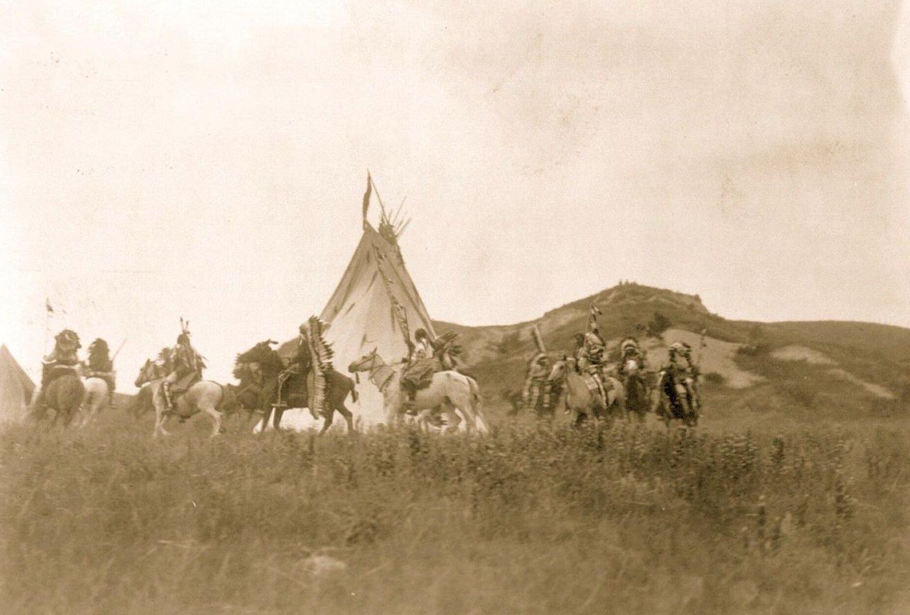 Several Dakota men on horseback riding in a circle around a tipi, 1907