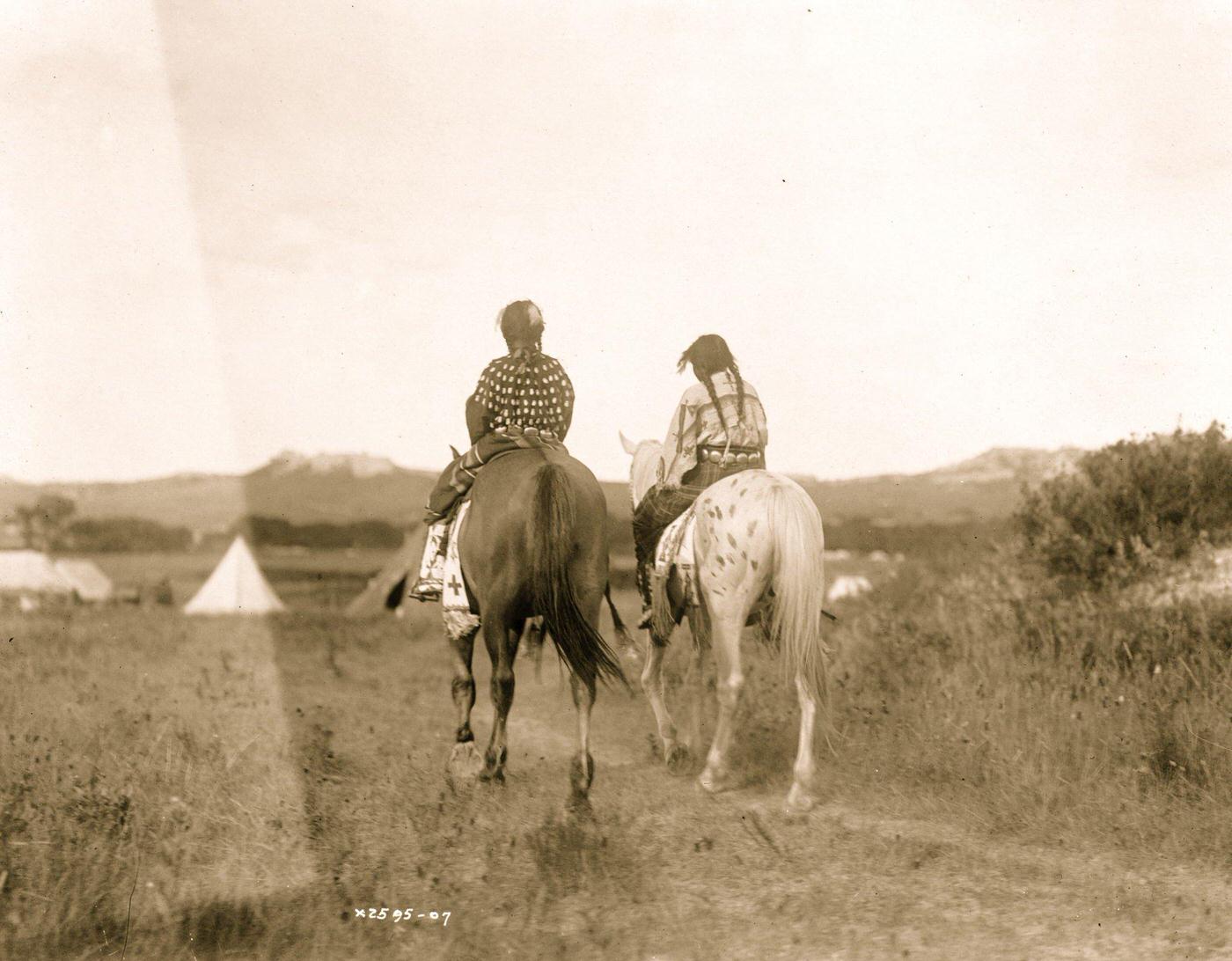 Two daughters of a chief on horseback, riding away from camera toward tents in background, 1907