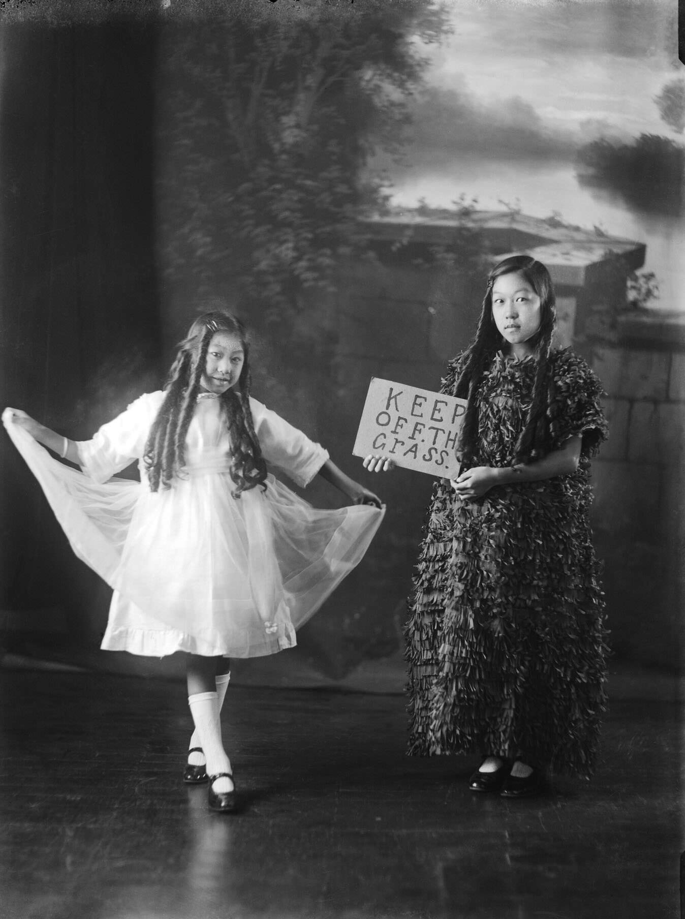 Two girls in fancy dress, one holding a sign saying 'Keep off the grass'