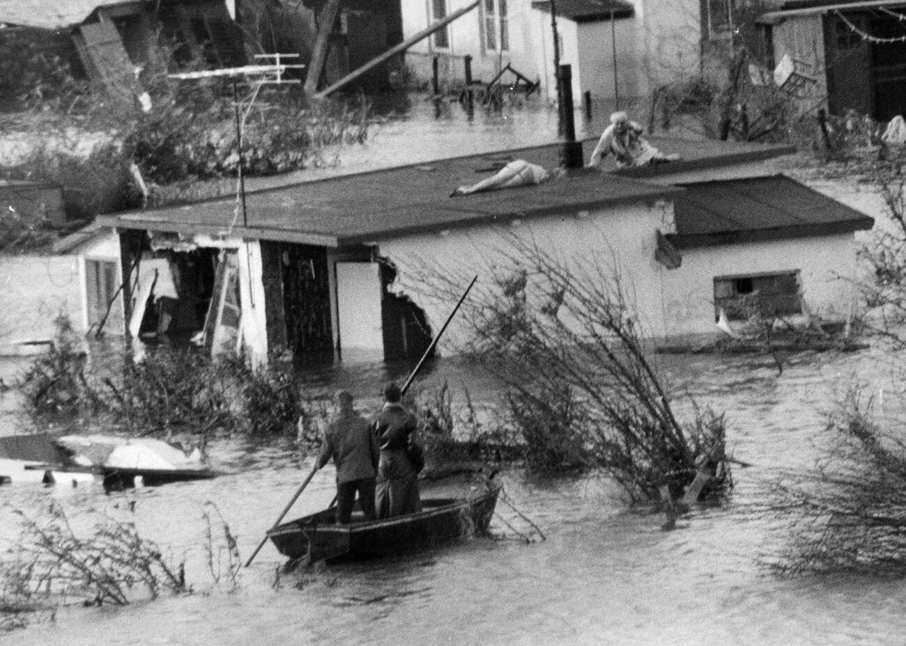 Man carrying the corpse of his wife on a roof after the North Sea flood of 1962 in Hamburg, West Germany, with victims and natural catastrophe in the background.