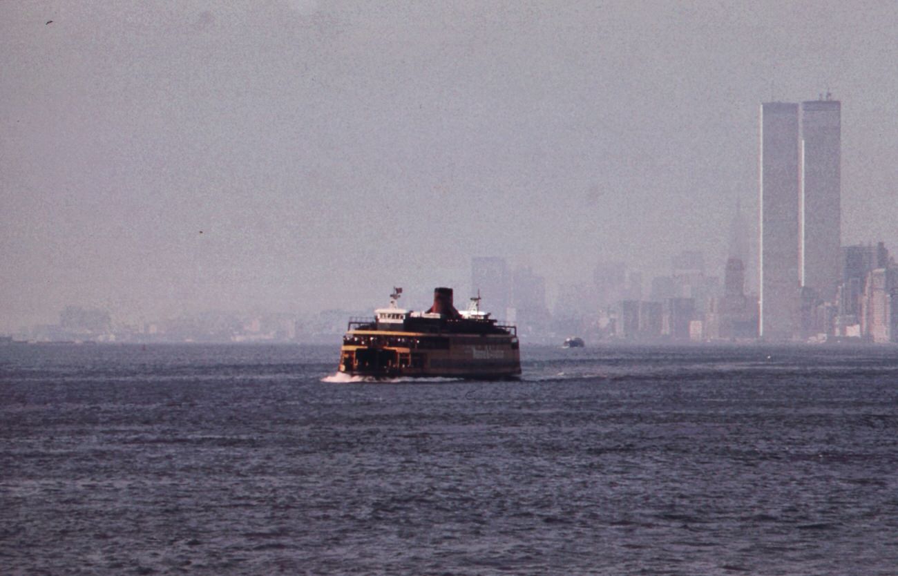 Staten island ferry with smog-obscured skyline of lower manhattan in background. On the right are the twin towers of the world trade center overlooking the hudson river, 1970s