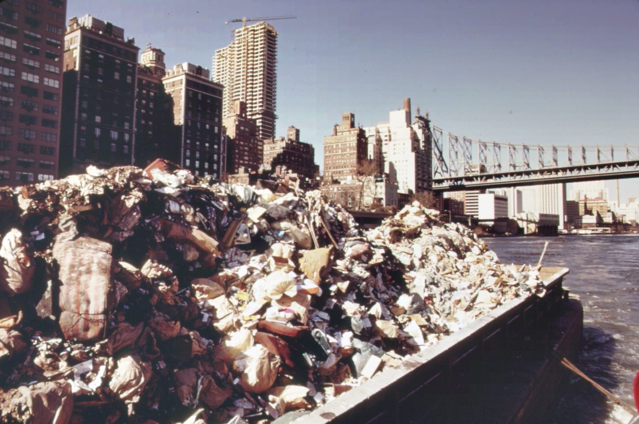 Part of the 26,000 tons of solid waste that new york city produces each day. Tugs tow heavily-laden barges down the east river to the overflowing staten island landfill, 1970s