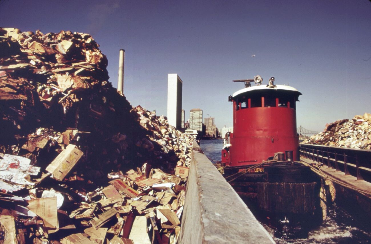 Tugboat herds two laden garbage scows down the east river from transfer point at 91st street. Destination is the staten island landfill, 1970s