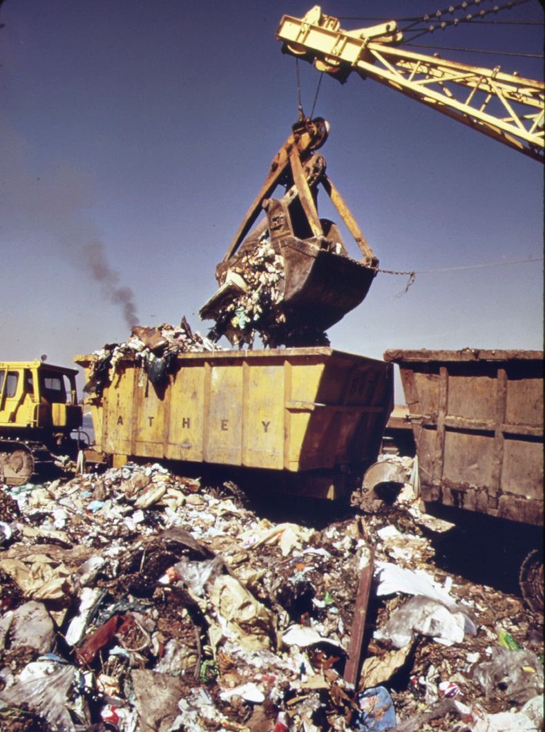 At staten island landfill steam shovel loads carts with garbage brought by barge from manhattan. Carts will haul refuse to outer edges of dumping area, 1970s