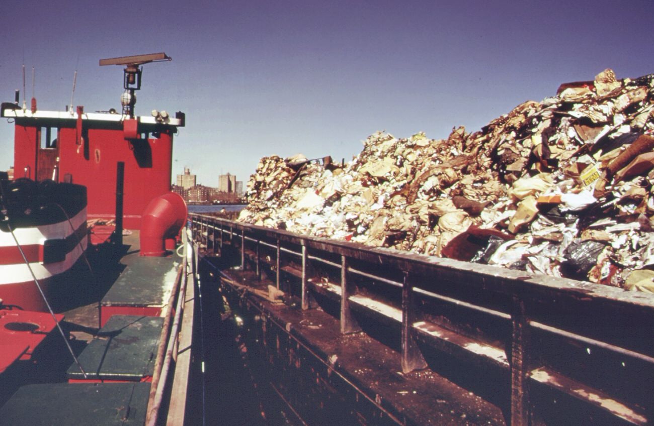 Tugboat tows heaped-up garbage scow from the 91st street marine transfer station down the east river. Garbage will be dumped at the staten island landfill, 1970s