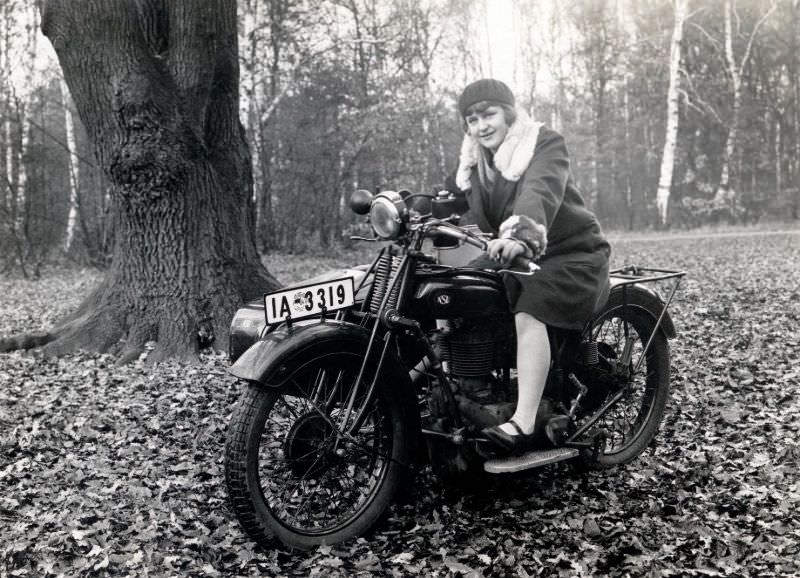 A young lady in a fur-trimmed coat posing in the saddle of an nsu 501 t combination in an autumnal forest. The bike is registered in the city of berlin, november 1929.