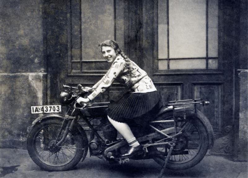 A cheerful young lady posing in the saddle of an ancient motorcycle in front of a double-leaf door. The words "1 january 1930" are handwritten on reverse. The bike is registered in the city of berlin.