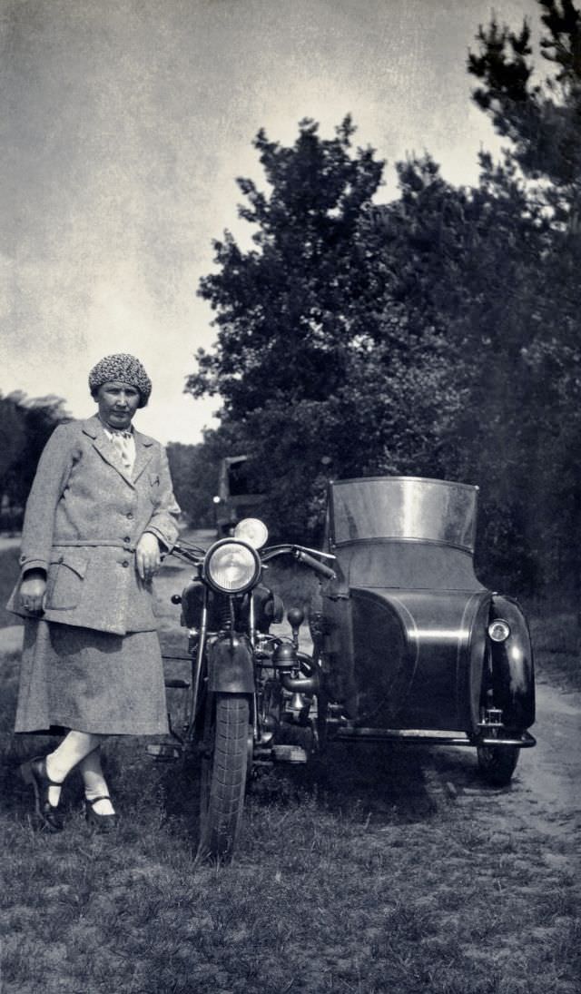 A stylish lady in a female suit posing with a sidecar motorcycle in the countryside, circa 1930s.